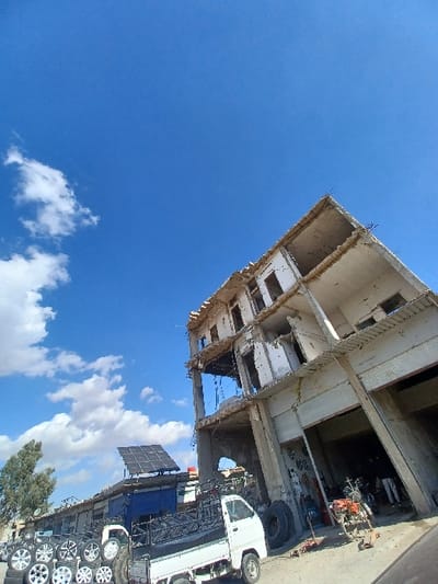 This image depicts an outdoor scene in Al-Qadam Municipality, Syria, under a clear blue sky with scattered cumulus clouds. The foreground and right side are dominated by a multi-story, partially constructed or damaged reinforced concrete building with exposed rebar and raw concrete surfaces, indicative of ongoing work or prior structural compromise. Its ground floor features large open bays. In the mid-ground, a white compact pickup truck is parked on an unpaved surface, its bed loaded with multiple stacks of silver vehicle wheels (rims and tires). To the left of the truck, a row of lower commercial structures is visible, one of which has an array of solar panels mounted on its roof. Two motorcycles are discernible near the entrance of the main concrete building, and several large tires lean against its pillars. A single bare tree stands on the far left. No discernible individuals or specific actions are evident within the frame.