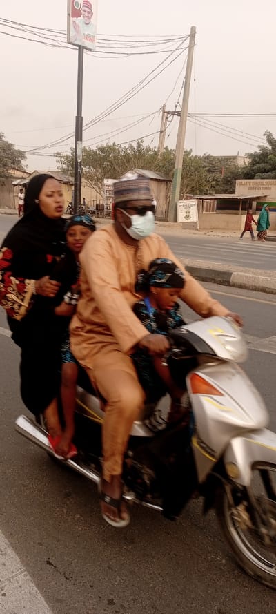 A man wearing a face mask and hat rides a silver motorcycle on a street. He is followed by two children and a woman. The woman wears a black hijab with floral patterns. A sign is visible on a pole in the background. Buildings and people walking can be seen in the distance on either side of the road.