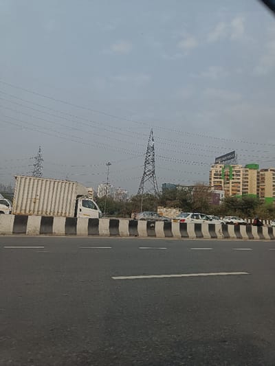 This image captures a busy urban landscape in Noida, India, under an overcast or hazy daytime sky. In the foreground, a multi-lane asphalt road with white markings is visible, separated by a concrete barrier painted with alternating black and white stripes.
On the left, a white delivery truck with a large, ribbed cargo container on its bed is moving on the main road. To the right of the barrier, several cars, mostly white sedans, appear to be either parked along the roadside or moving slowly in a congested lane.
Above the scene, numerous power lines traverse the sky, supported by two prominent, tall electrical transmission towers (pylons) made of steel lattice structures. Beyond the roadside vegetation and these towers, the background reveals a dense cluster of multi-story buildings. A particularly noticeable building features light-colored facades with green accents and has a large billboard mounted on its rooftop. The text "GAUR CITY CENTRE" is clearly visible on this billboard. The distant buildings fade into the hazy atmosphere, completing the view of a bustling Indian city.