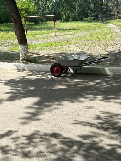 An outdoor scene in Angarsk, Russia, shows a wheelbarrow with two red wheels and green handles parked on asphalt next to a concrete curb. Two wooden poles lean against a tree trunk painted white near the base. Behind the wheelbarrow is a grassy area with a red metal soccer goal. The background features trees and a multi-story building. Sunlight casts shadows of leaves onto the asphalt in the foreground.
