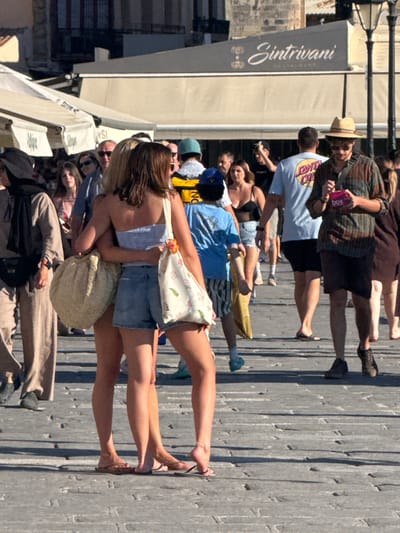 In Chania, Greece, on a paved promenade, two young women are embracing. The woman on the left has blonde hair and is wearing a light blue strapless top and denim shorts. The woman on the right has brown hair and is wearing a white and floral print top. She is also wearing denim shorts. Both women are carrying large tote bags. They are surrounded by a crowd of people walking in both directions. The background shows buildings with awnings and shops. In the distance, a sign reads "Sintrivani Restaurant". The scene is brightly lit, suggesting it is daytime.