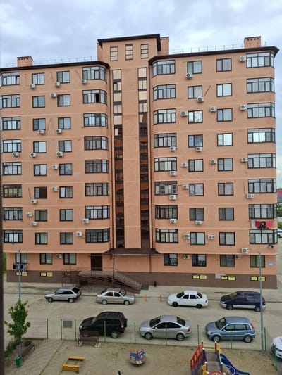 This is a medium-angle, full-frame photograph taken from an elevated position, looking down at a modern, multi-story apartment building and its surrounding area in Yablonovskiy, Russia. The building, constructed primarily of light brown brick, has a complex, multi-faceted design with numerous windows and air conditioning units visible on its facade. The sky is overcast with gray clouds.
In front of the building, on a paved surface, are several parked cars. From left to right, there is a silver hatchback, a silver sedan, a white sedan, a dark blue minivan, a silver sedan, and a blue minivan. Further to the right, a white car is partially visible. Beyond the paved area, there is a fenced-off playground with a slide and sandbox. A lone tree stands to the left of the building, near a green trash can and a park bench. The area appears to be a residential complex. No people are visible in the frame. The overall impression is one of an urban residential setting under a cloudy sky.