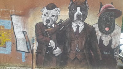 This is a photograph of a mural painted on a textured, weathered wall, possibly in Kryzhanivka, Ukraine, given the context. The mural depicts three anthropomorphic dogs dressed in formal attire.

On the left, a light-colored dog, possibly a bulldog, wears a brown suit, a white shirt, a bow tie, and a dark flat cap. Its left paw rests on a chain or pocket watch chain.

In the center, a black and white pit bull-type dog is dressed in a brown suit, a white shirt, and a dark tie with a subtle pattern. It wears a similar dark flat cap, tilted slightly. This dog has its right arm resting on the shoulder of the dog to its left, and it appears to be holding a dark object, possibly a pipe or a rolled document, across its chest. A pocket watch chain with a charm is visible on its vest.

On the right, a black Scottish terrier-type dog is depicted wearing a pink-brimmed hat. It has a white shirt and appears to be wearing a vest, though the details are less clear due to the dog's dark fur and the mural's texture. Its mouth is open, revealing a pink tongue.

The background of the mural is a mottled orange and yellow, with some graffiti elements visible on the left side, including abstract shapes in blue and yellow. A vertical metal pole with a greenish top is situated in front of the mural on the left side. To the far right, the edge of a white vehicle and a silver car are partially visible, suggesting an urban or semi-urban setting. The overall style of the mural is realistic, with detailed rendering of the dogs' fur and clothing.