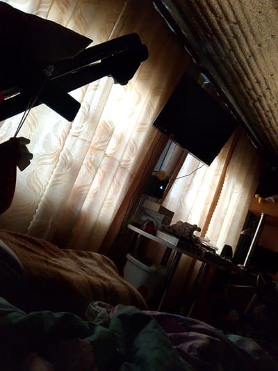 The image depicts a cluttered bedroom interior, likely in Savino, Russia. The scene is dominated by soft, patterned curtains illuminated by natural light filtering in from a window. In the foreground, a bed covered with blankets is visible. To the right, a metal-framed table holds various items, including a television set mounted on the wall above it. Further back, what appears to be a sloped ceiling constructed of corrugated material is present. The overall impression is of a lived-in, somewhat disorganized domestic space.