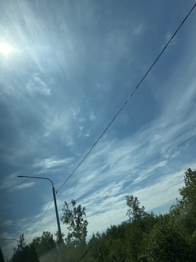The image captures a bright, overcast sky with wispy clouds. A power line stretches diagonally across the frame. On the left side, a street lamp is visible, with another one partially visible further in the background. The lower portion of the image shows a dense line of green trees, indicating a natural or semi-natural environment on the outskirts of a populated area. The sun, partially obscured by clouds, is visible in the upper left corner, casting a bright glare. Based on the context of the city of Aur, Russia, this scene is likely a roadside or park area within or bordering the city, given the presence of street lamps and overhead wires.