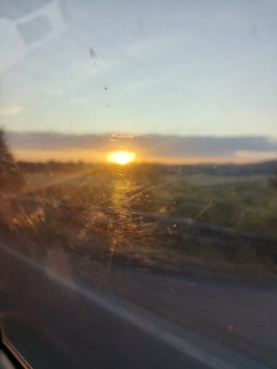 The image depicts a hazy sunset viewed through a dirty window, likely from inside a vehicle. The sun, a bright orange orb, is partially obscured by clouds and casts a warm glow across the landscape. The foreground is dominated by the reflection and smudges on the windowpane, which distort the view of the distant, blurred scenery. The scene is devoid of any people or explicit city structures, suggesting a rural or semi-rural setting on the outskirts of Radobyčice, Czechia. The overall impression is one of a fleeting, melancholic moment captured during twilight.