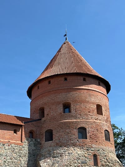 The image shows a close-up view of a circular red brick tower with a conical red tile roof, a prominent feature of Trakai Island Castle in Trakai, Lithuania. The tower has several arched windows with dark panes and is built on a base of large, rough stones. A smaller, attached brick building with a red tile roof and similar stone base is visible to the left. The sky is clear and blue, with a small black flag at the pinnacle of the tower and two birds flying in the distance. The overall scene depicts a historic defensive structure against a bright, sunny sky.