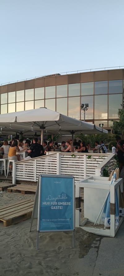 The image depicts an outdoor dining area at a beach establishment, likely located along the Danube River in Linz, Austria, given the "Sandburg" branding on a sign. The scene is set during the late afternoon or early evening, with soft, warm light reflecting off a modern building's large windows in the background.
In the foreground, a blue A-frame sign displays text in German, indicating that "Liegestühle (loungers) are ONLY FOR OUR GUESTS!" and also requesting guests to return their "Chill-Session" items. The sign is partially submerged in wet sand. To the right of the sign, a white wooden structure, possibly a station for storing beach chairs or umbrellas, is visible. Several blue and white striped beach umbrellas or towels are stored within this structure.
Behind the sign and storage unit, a white wooden railing encloses a dining area. Several people are seated at white tables under large white umbrellas, enjoying food and drinks. They appear to be engaged in conversation and socializing. The ground surface is a mix of sand and some wooden planks, suggesting a beach or waterfront setting. The overall atmosphere is relaxed and social, characteristic of a summer evening at a riverside establishment.