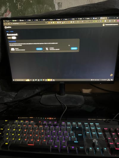 The image shows a computer monitor displaying a webpage with the title "Account." The webpage is a social platform's account linking settings page, displaying options for linking Discord and Twitter accounts. The monitor is black and has the brand name "AOC" printed on the lower right side of the screen. It is sitting on a desk with a black, backlit keyboard in front of it. The keyboard is slightly dusted. The image is taken from a low angle, with the focus on the monitor and keyboard. The surrounding environment is dimly lit. The location is the city of Kecamatan Waru in Indonesia, based on the provided coordinates.