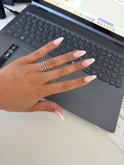 An overhead shot shows a woman's left hand resting on a grey laptop, with a paper on the right side of the laptop. The woman's nails are painted with a French manicure, and she is wearing a silver ring with a geometric design. The laptop is open and appears to be in use, with the screen visible. The background is out of focus but suggests an indoor environment, possibly an office or study. No specific location in Les Arcs, France, can be determined from the image.