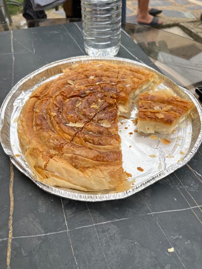 A partially eaten, spiral-shaped pastry with a golden-brown, flaky crust is presented in a round aluminum foil pan on a dark, speckled table. The pastry is sliced into several portions, with one piece removed and placed to the side, revealing a white, creamy filling. Small, light-colored flakes, likely from the crust or topping, are scattered around the pan. Behind the pastry, a clear plastic bottle of water is visible. In the background, out of focus, are the legs of two individuals wearing sandals, standing on a light-colored, textured surface that appears to be an outdoor patio or walkway. The environment suggests an informal outdoor setting, possibly a backyard or terrace, in Stains, France, given the information provided.
