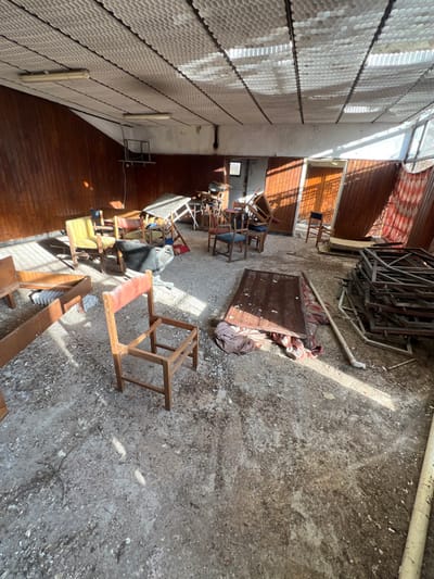 The image displays a large, disused indoor hall located in Stoianovo, Bulgaria. The floor is concrete, heavily soiled with dust, dirt, and fragmented debris. Natural sunlight streams from unseen windows on the right, casting distinct bright patterns across the floor.
The lower two-thirds of the walls are paneled with dark brown wood, while the upper portion is a discolored, light-colored painted surface. The ceiling features a textured, light-colored hexagonal or honeycomb pattern, with exposed black structural tracks and two non-illuminated fluorescent light fixtures.
Numerous pieces of furniture are scattered throughout the room in disarray. In the foreground, slightly left of center, stands a wooden chair frame with an incomplete seat and a faded red upholstered backrest. To its left, a disassembled brown wooden shelving unit lies on its side, revealing a stack of white, brick-like objects.
In the mid-ground, several chairs are visible, including a yellow upholstered armchair, a black object resembling a damaged office chair or bag, and multiple wooden chairs, some with blue upholstery, stacked or overturned. Various tables, some folded or propped against other items, are present. A flat, rectangular wooden panel or tabletop lies on the floor to the right of the incomplete chair, with dark fabric partially visible beneath it.
On the far right, a large pile of metal frames or disassembled structural components is stacked on the floor. A long, white cylindrical object, possibly a pipe or rolled material, rests on the floor nearby. Two wooden doors are visible on the right wall, one slightly ajar, and a section of red and white patterned fabric is draped over an unseen structure near the far right corner. A small, square opening is visible on the far-left wall. No individuals are present in the scene.