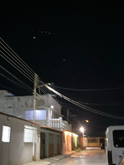 This image depicts a nighttime street scene in La Cruz del Pastel, Venezuela. The sky is dark, showing several faint, elongated, light-blue or greenish illuminations, including two distinct crescent-shaped blue lights in the upper quadrants and a cluster of four smaller, fainter lights in the upper central area. Overhead, numerous utility lines traverse the frame, originating from a prominent utility pole positioned left of center.

On the left side of the street, a single-story building with a light-colored facade features barred windows. Adjacent to it, a two-story building with a light-toned exterior presents a balcony with a white balustrade on the second level and a window covered by vertical white slats. The street, which appears paved, extends into the distance, illuminated by multiple streetlights. One bright streetlight is mounted on the utility pole, casting white light, while other streetlights further down emit warmer, orange light. Buildings lining the right side of the street also display illuminated doorways or windows with warm light. A white vehicle, possibly a van, is partially visible on the far right. No individuals are present, and no active interactions or discernible actions are observed.