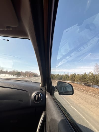 The image shows a view from inside a car, likely the driver's seat, looking out the passenger side window. The car is on a road with trees lining one side. There appears to be some snow on the ground. The side mirror is visible. The dashboard is black and has a circular air vent.
I am unable to identify the location as Pakli, Russia as it is not visible in this image.