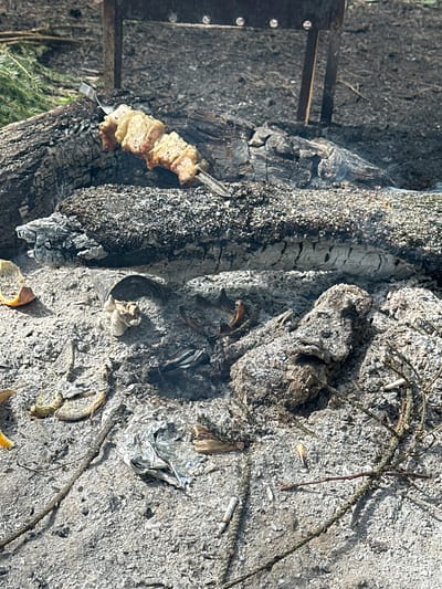 The scene depicts the aftermath of a fire. Ash and charred wood dominate the view. A metal skewer with partially cooked food rests near the burnt remains. Cigarette butts and debris are scattered among the ashes. The image suggests an outdoor setting, possibly in a rural or recreational area within or near Юзуфоўскі сельскі Савет, Belarus. No people are visible.