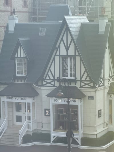 The image displays a prominent Louis Vuitton boutique located at the corner of Rue Eugène Colas in Deauville, France. The building features a distinctive Anglo-Norman or Tudor Revival architectural style, characterized by a white facade with dark timber framing, multiple gables, and steeply pitched dark slate roofs. Two prominent white chimneys are visible on the roofline.
The ground floor of the building houses the Louis Vuitton store. To the left, a white-railed staircase leads up to the main entrance, above which "LOUIS VUITTON" is displayed in white lettering against a dark background. To the right of the entrance, a large display window showcases various Louis Vuitton merchandise, including handbags and accessories, arranged on shelves. The brand's "LV" monogram logo is affixed to the facade both next to the display window and further to the right. A black, ornate street lamp with a globe fixture stands directly in front of the display window.
In the lower right portion of the image, one individual is observed from behind, standing on the pavement in front of the display window, seemingly looking into the store. The background reveals other structures, including a lighter-colored building with some visible brickwork and, further back, scaffolding indicating ongoing construction or renovation. The street surface in the foreground appears damp or reflective, suggesting a recent rain or overcast conditions.