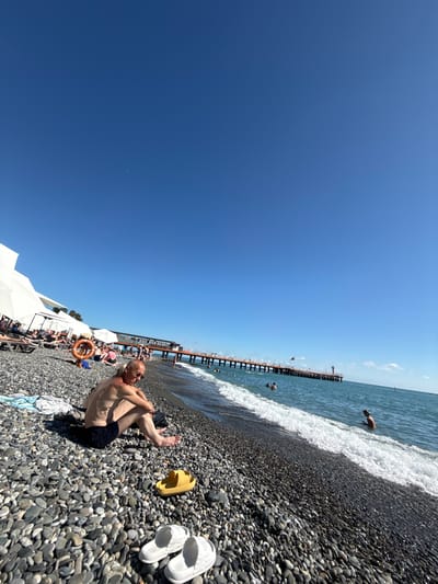 A wide-angle, eye-level shot captures a sunny day at a pebble beach in Adlerskiy, Russia. In the foreground, an older man with sunglasses and dark swim trunks sits on the pebbles, looking down with his arms resting on his knees. Next to him are a bright yellow sandal and a pair of white flip-flops. In the mid-ground, a colorful orange life preserver hangs on a stand. White canopies and lounge chairs are set up further down the beach, with various people seated and reclining. A long pier with structures and a red lighthouse extends into the blue sea. Several people are visible in the water, with waves lapping at the shore. The sky is a clear, vibrant blue with a faint crescent moon visible. The overall scene depicts a relaxing day at the beach.