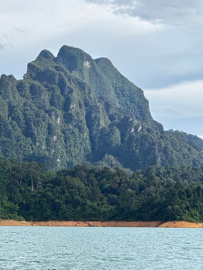 A wide, horizontal, eye-level shot captures a serene natural landscape in Khao Phang, Thailand. In the foreground, rippling, turquoise water fills the bottom third of the frame, with a suggestion of dried-out tree stumps visible just at the water's edge, protruding from a reddish-brown embankment. This embankment forms a gentle curve along the water.
Dominating the mid-ground and background is a massive, verdant mountain range covered in dense, dark green foliage. The mountain peaks are rugged and uneven, with visible rock faces interspersed with the thick vegetation. The overall impression is one of lush, untouched wilderness. The sky above is overcast with light gray clouds, casting a soft, diffused light over the entire scene. The image is devoid of any human presence or man-made structures, emphasizing the raw beauty of the natural environment.
