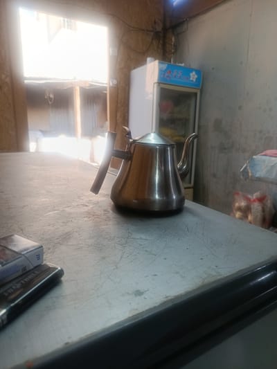 The image depicts an interior scene, likely a small shop or dwelling, located in Kadji Say, Kyrgyzstan. In the foreground, a metallic teapot with a wooden handle rests on a scratched, grey surface, possibly a countertop. To the left of the teapot, a pack of cigarettes and a lighter are visible. In the background, a white refrigerator with a blue sign featuring Chinese characters and a star is present. Behind that, a brightly lit opening, possibly a window or doorway, reveals an outdoor area with corrugated metal structures. The walls of the interior appear to be made of plywood or a similar material. The scene is dimly lit, with sunlight streaming in from the opening in the background. There are no people visible in the image.