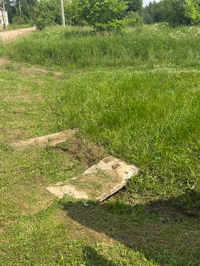 The image depicts an outdoor scene in the village of Borodulino, Russia, characterized by lush green grass and a dirt path. Two concrete slabs are partially visible, positioned on the ground and covered by grass. One slab is oriented horizontally in the lower part of the frame, while another, slightly higher and to the left, is angled diagonally. A small patch of disturbed earth is present between these two slabs. The surrounding environment consists of tall grass, with trees and foliage visible in the background, suggesting a rural or semi-rural setting. The lighting indicates it is daytime, with shadows cast on the grass. There are no people or animals present in the image.