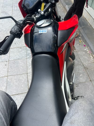 The image captures a close-up, high-angle perspective of a red and black motorcycle parked on a paved sidewalk. The motorcycle's seat is black and textured. On the fuel tank, two stickers are visible: one with a blue circular icon and another with a white circular icon featuring a blue motorcycle and the number "90+". The handlebars, with a black grip and control switches, are prominent in the upper left. Part of a person's legs, clad in grey jeans, is visible in the foreground on the left and right. The background shows a person in dark clothing standing behind the motorcycle. The sidewalk is composed of grey square tiles. The setting is the city of Hà Giang 2 Ward, Vietnam.