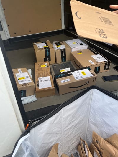 The image depicts the interior rear cargo area of a delivery vehicle, likely a van, filled with numerous packages. The scene is located in Seligenstadt, Germany, as evidenced by a shipping label on a visible package that reads "Kortenbacher Weg 14, 63584 Seligenstadt, Germany."

A person's right hand is visible in the upper right quadrant, holding an open brown Amazon Prime padded envelope marked "PM3" and bearing the Amazon smile logo. Below this, the cargo area floor is covered with various Amazon Prime cardboard boxes and brown paper mailers. The boxes are in different sizes and orientations, some taped with "amazon prime" branded tape. Several packages have yellow labels with codes such as "D7", "D77 3E", "PRG2", and "CYCLE 1". One large rectangular box on the right is labeled "BONTEC" and "Model/Modell: MF1039-00-1". Another white rectangular box is visible with "amazon.com" printed on it.

On the dark-colored floor of the van, to the right of the packages, are two mobile phones; one black and one with a dark green or gold frame and black screen.

In the foreground, a large, rectangular, white insulated delivery bag or container is partially open, its white interior lining visible and slightly wrinkled. The opening is secured by an orange clip attached to the vehicle's frame. The bag contains additional brown paper Amazon Prime envelopes, stacked upright.

The interior walls of the vehicle are paneled with light brown, possibly fiberboard or particle board, secured by visible black bolts. The floor is a dark, uniform surface.