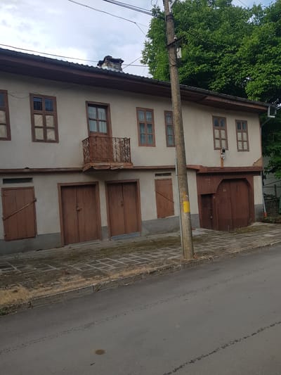 A slightly angled, medium shot captures the facade of an aged, two-story building with a light-colored stucco exterior and a dark brown tiled roof in the city of Lomets, Bulgaria. The building exhibits signs of wear, with some crumbling plaster visible near the roofline.

The ground floor features several large, dark brown wooden doors, suggesting they may be garages or workshops. To the left, a narrower, similar wooden door is set into the wall. Above these doors, the second story has a row of four evenly spaced, multi-paned windows with dark brown frames. A small, ornate, dark brown metal balcony juts out from the second story, situated between the second and third windows from the left. A concrete sidewalk with a patterned tile surface runs along the front of the building, bordered by a strip of dry grass.

A thick, concrete utility pole stands prominently in front of the building, slightly to the right of center, supporting numerous electrical wires. A large, mature tree with dense green foliage dominates the upper right portion of the image, casting shadows on the building and obscuring part of the sky, which is overcast with soft clouds. The foreground shows a gray asphalt road, with faint tire tracks and a small, dark stain visible. The overall impression is one of a rural or older urban setting with structures that have seen considerable use over time.
