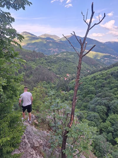 A man stands on a rocky precipice, observed from a slightly elevated position. He wears a white shirt, dark shorts, and white shoes. The background reveals a mountainous landscape densely covered in green trees. A bare, partially broken tree branch extends across the scene. The sky is partially visible, with patches of blue and white clouds. The location is identified as Zasele, Bulgaria.
