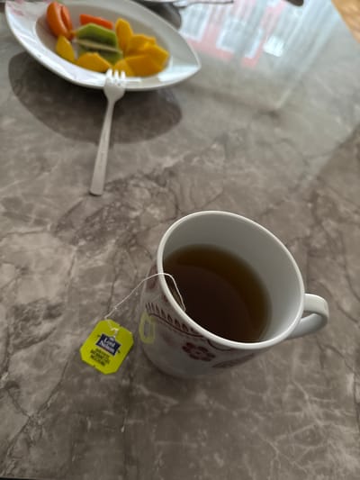 The image depicts a mug of tea and a plate of fruit on a marble-patterned surface. The mug is white with a brown floral design and a yellow tea bag tag with "Lord Nelson" printed on it. The fruit plate contains slices of mango, kiwi, and orange. A white plastic fork rests on the plate. The environment suggests an indoor setting, potentially a kitchen or dining area. There's nothing in the image to indicate Basel, Switzerland.