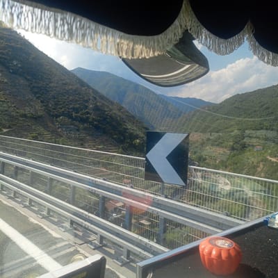 The image appears to be taken from inside a vehicle traveling on a highway overpass. In the foreground, there is a dark-colored interior trim with white fringe hanging down, obscuring the top part of the view. Below this, a shiny surface, possibly a dashboard or interior panel, reflects the sky and the road ahead.

The road itself has white lane markings and a gray asphalt surface. Guardrails made of metal poles and horizontal beams run along the side of the overpass.

Beyond the guardrails, a steep, mountainous landscape is visible. The hills are covered with dense vegetation, appearing as a mosaic of green and brown. Terraced slopes with vegetation are carved into the hillsides, suggesting agricultural use. Several small buildings or houses are scattered throughout the landscape.

A prominent black and white chevron sign, indicating a sharp left turn, is mounted on a pole to the right of the road.

In the far distance, more mountains are visible, fading into a hazy blue. The sky is partly cloudy with bright sunlight.

On the right side of the image, on the surface below the guardrail, a small, orange, pumpkin-shaped object with a label on top is visible.

While the specific location of Taggia, Italy is mentioned, the visual elements themselves do not provide direct confirmation of this location. The landscape is consistent with mountainous regions of Italy.