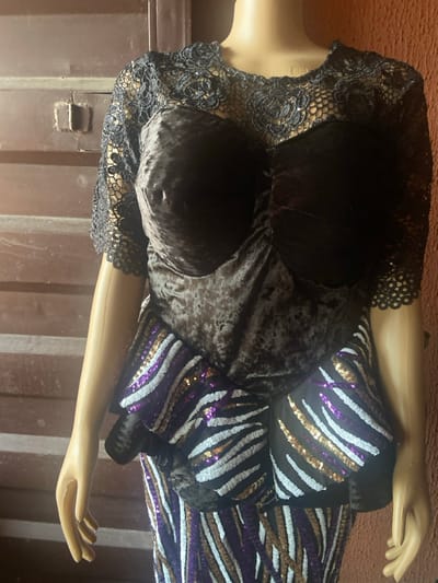 A mannequin displays a dark velvet and sequined dress. The bodice features a dark velvet bra-like top over a crushed velvet midsection, trimmed with black lace. The dress's skirt is adorned with tiger-like stripes of sequins in white, purple, and gold. The mannequin's painted arms are extended, and the background appears to be a brown metal container.