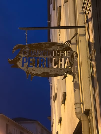 The image shows a weathered, pig-shaped sign with the words "CHARCUTERIE PETRIUCHA" cut out in blue letters. The sign is hanging from a chain attached to a metal bracket mounted on the side of a building. A fork graphic is also integrated into the sign. The building appears to be made of stucco and has several windows. A downspout is visible running vertically along the wall. The scene is illuminated by artificial light, casting shadows. The background is a deep blue night sky. The sign is located on a street in Vienne, France, as indicated by visual context and common signage for charcuterie shops in the region. There are no people or animals visible in the image.