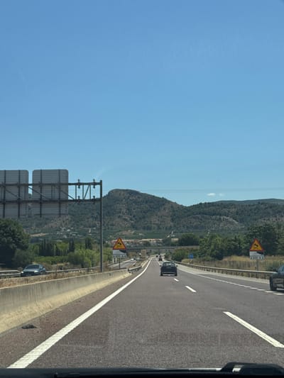 The image shows a view from a vehicle traveling on a highway. The highway has multiple lanes separated by white lines and a concrete barrier. In the foreground, the asphalt road is visible with white lane markings. In the middle ground, there are other vehicles on the road, including a dark-colored car in the right lane and a lighter-colored car further ahead in the left lane. There are also traffic signs visible. To the left of the highway, there is a concrete barrier and some vegetation. In the background, there are rolling hills covered with trees and some buildings can be seen in the distance. The sky is clear and blue. The scene is consistent with a highway in a rural or semi-rural area, possibly the outskirts of Canals, Spain, given the terrain and vegetation.