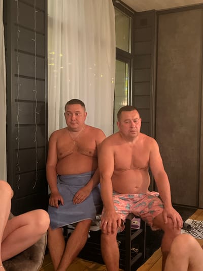 The image depicts two men in what appears to be a sauna or relaxation area. One man is wearing pink shorts with a tropical print and is sitting on a black shelf or bench. The second man is wearing a light blue towel wrapped around his waist. A third person, only visible from the knees down, is near the bottom of the frame. The background suggests an indoor setting with a window partially covered by white curtains with string lights. The location cannot be verified to be Новая Чернушка, Russia, based on the provided image.