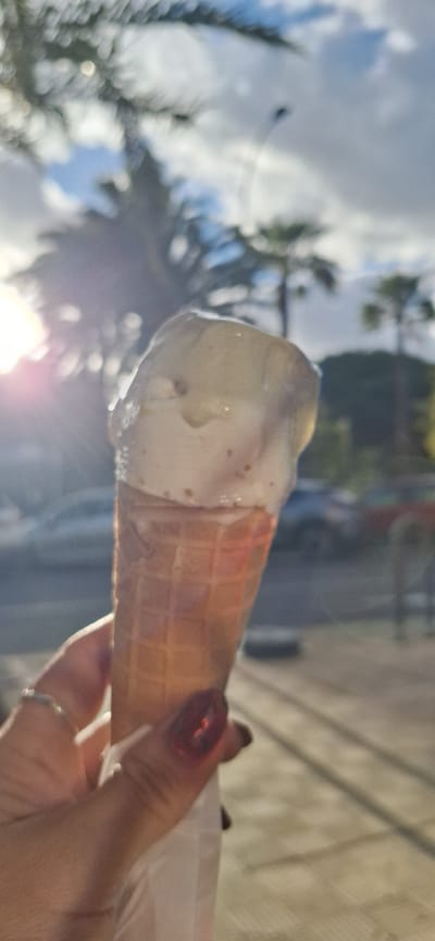 A photograph shows an ice cream cone held in a person's hand. The ice cream is light-colored and appears to be melting slightly. The cone has a waffle pattern and is partially wrapped in paper. The person's hand is visible, with painted fingernails and a ring on one finger. The background is out of focus but appears to be an outdoor scene with trees and a sky with clouds. The image's angle suggests it was taken by the person holding the ice cream. The location is Santa Cruz De Tenerife, Spain.