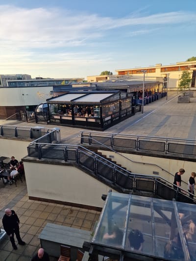 The image shows an outdoor area of a restaurant or bar in Durham, United Kingdom. The camera angle is elevated, possibly from a higher floor or rooftop. The sky is light blue with scattered wispy clouds.
The restaurant has a covered outdoor seating area with many customers sitting at tables. The structure is modern with glass and metal elements, likely a popular dining spot.
Staircases with metal railings lead up to the restaurant, suggesting a split-level layout. A partially visible outdoor seating area with tables and chairs is located on a lower level. People are seated and interacting at the tables.
The architecture includes modern buildings and structures. The paving stones are light-colored and create a pedestrian-friendly zone. In the lower right corner, there is a glimpse of an outdoor table and people, with one person appearing to walk away.