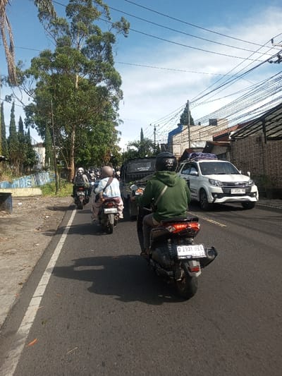 The image captures a street scene dominated by vehicular traffic under a clear blue sky with scattered white clouds. The perspective is from behind a black Yamaha NMAX-type scooter, displaying the Indonesian license plate "D 2931 BEE" with a "01-20" validity tag, indicating a West Java (Bandung region) registration.

Ahead of the camera, a line of motorcycles is observed traveling in the same direction. The closest motorcycle in this line is a lighter-colored scooter, bearing the Indonesian license plate "D 6724 AG". All visible motorcyclists are wearing helmets. To the right of this scooter, a dark-colored pickup truck is partially visible, also moving away from the camera.

On the right side of the road, a white SUV, consistent with a Toyota Rush or Daihatsu Terios, is present, possibly moving in the opposite direction or stationary. It features a roof rack with items secured by a blue tarp. Its license plate, "F 1726 WA", also indicates a West Java (Bogor/Sukabumi/Cianjur region) registration.

The asphalt road surface features a solid white line on the left, delineating a shoulder or a separate lane. Distinct shadows from vehicles and trees are cast onto the road, indicating bright, direct sunlight. The left side of the road is characterized by a blue-painted concrete barrier with graffiti, a grassy verge, and a dense line of trees, including tall, broadleaf specimens and slender conifers. The right side of the road is lined with buildings of varying designs, with numerous overhead utility wires spanning across the sky.