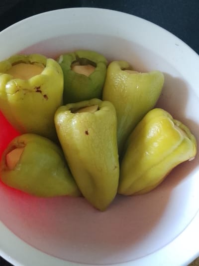 A close-up, overhead shot shows several light green bell peppers, stuffed with a light-colored filling, resting in a white bowl. The peppers are arranged closely together, with their tops opened to reveal the filling. The bowl is partially filled with a pinkish liquid, and the background is dark and out of focus. The scene is likely indoors, possibly a kitchen, and given the context, it is in the city of Kerben, Kyrgyzstan. There are no people visible in the image.