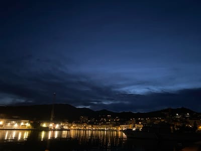 A nighttime view of a harbor town reflects in calm water. Illuminated buildings and streetlights line the shore, casting golden reflections. A dark silhouette of a ship sits in the foreground. A tall tower rises behind the town. Dark mountains serve as a backdrop beneath a deep blue sky filled with wispy clouds. A few faint stars are visible above.