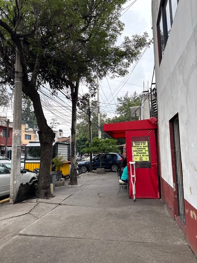 The image shows a sidewalk in Tizapan, Mexico, with a building on the right and trees and a street on the left. A red awning covers a storefront or entrance. A yellow sign on the red awning reads "DESAYUNOS, CAFÉ O TÉ, FRUTA O JUGO, HORARIO DE ATENCIÓN 8:30 A 12:30". A person wearing a teal shirt is seated at a table next to the red awning, and a pair of crutches leans against the awning. The sidewalk is cracked and uneven. There are parked cars visible on the street.