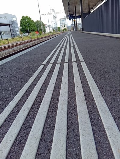 The image shows a close-up view of a train station platform in Aarburg, Switzerland. The foreground is dominated by tactile paving, consisting of parallel white strips set into a dark grey asphalt surface. These strips are likely intended to guide visually impaired pedestrians. Beyond the paving, a set of train tracks is visible, bordered by a metal fence. In the mid-ground, a modern building with grey cladding and windows is partially visible, along with trees and overhead power lines for the trains. Further in the distance, more buildings and structures of the town can be seen under an overcast sky. The scene is a typical urban railway environment.