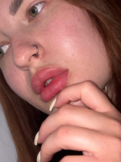 A close-up image shows a person's face with a nose piercing. The person is pulling their lip. The background is out of focus. The person has light skin, light brown hair, and hazel eyes. The fingernails are painted a light color. There is no information available to determine the location as Port-Katon, Russia.