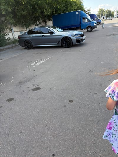 This media file depicts a street scene in Tambov, Russia, likely during the daytime with clear weather. The primary subjects are several vehicles parked on an asphalt road. In the foreground, a sleek, gray sedan with tinted windows and black rims is prominently featured. Behind it, a blue truck with a covered cargo area is visible, followed by a smaller blue truck with a lighter blue cab.
The scene appears to be a parking area or a street with multiple vehicles. Trees line the left side of the frame, suggesting an outdoor setting with some greenery. A partial view of a person wearing a floral-patterned garment is visible at the top right of the image, with their arm and shoulder in frame. The person's presence suggests a possible social gathering or everyday activity. The overall impression is one of a casual outdoor environment, possibly a parking lot or a quiet street. No explicit activity or event is clearly depicted, beyond the vehicles being parked. The lighting suggests it is daylight, and the asphalt surface and clear sky hint at fair weather.