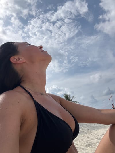 A low-angle shot reveals a woman with dark hair, wearing a black bikini, lying on a beach. A thin silver necklace rests on her chest. Above, a sky filled with white clouds is visible, along with a palm tree and a distant object resembling a volleyball net. The sandy beach can be seen to the right of the woman. The scene appears to be set in Baan Chaloklum, Thailand.