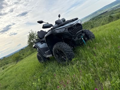 The image shows a gray four-wheel ATV parked on a grassy hillside. The ATV is facing forward, and the ground slopes down from the right to the left. The sky is cloudy. In the distance, there is a town in a valley, trees, and hills. Based on the location, the town in the distance is likely Blagoevo, Bulgaria. There are purple flowers interspersed throughout the grass.