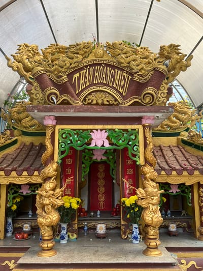 This image depicts a highly ornate, multi-tiered altar structure identified by the prominent gold Vietnamese inscription "THẦN HOÀNG MIẾU" on a red plaque at its apex, located in Đăk Mar, Vietnam. The structure is predominantly gold, red, and brown, featuring extensive sculptural details.
The uppermost section is adorned with multiple golden dragon figures intricately carved and facing outward. Below this, a reddish-brown tiled roof section with textured details is visible. The central frontal area of the altar is an open recess framed by gold and green decorative elements. A large pink lotus motif is centered above the inner opening, which features red panels with vertical gold Vietnamese script.
Two golden dragon sculptures serve as pillars flanking the main altar entrance, resting on the lower platform. On the altar platform itself, various items are arranged symmetrically: several vases containing yellow flowers (chrysanthemums), bowls of red fruits (appearing to be apples or pomegranates), and multiple smaller ceramic or porcelain vessels, some with blue and white patterns, potentially functioning as incense burners or offering bowls. Small, unlit candles are also present on the platform.
The entire altar is situated beneath a translucent, arched corrugated roof structure supported by metal framework, providing shelter. Green foliage is visible in the background, suggesting an outdoor or semi-enclosed garden setting. No individuals or active interactions are visible within the frame. The materials show slight signs of weathering and environmental exposure, such as minor discoloration on the roof tiles.