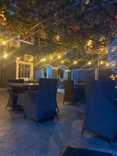 This image depicts an illuminated outdoor dining or lounge area at night, located in Igbessa, Nigeria. The scene is devoid of people. Overhead, a dark metal trellis structure is adorned with artificial green and autumnal-colored foliage, from which multiple strings of warm-toned bare bulb lights are suspended and illuminated. The ground is a dark, possibly textured concrete surface.
In the mid-ground, a square table with a glass top is centered, surrounded by four dark brown or black wicker-style chairs. Additional tables and chairs of similar design are visible further into the background, arranged on the concrete floor.
In the background, a dark building wall is visible on the left, featuring a white-framed window with horizontal blinds and two white wall-mounted air conditioning units. To the right of this, two arched structures, potentially entrances, are illuminated with a blue light from within, with a sign reading "BAR" visible above one arch. A large, light-colored industrial fan's protective cage is partially visible on the far right.