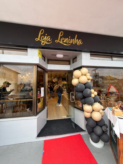 The image depicts the exterior and partial interior of a commercial storefront identified as "Loja Leninha" in Felgueiras, Portugal.
The store's facade features a black sign with "Loja Leninha" inscribed in gold script. Below the sign, a central glass door serves as the entrance, flanked by large glass display windows. A red carpet extends from the threshold onto the gray concrete pavement outside, indicative of a special event or opening. A large, organic-style balloon garland, composed of beige and black balloons of various sizes, is positioned vertically to the right of the entrance, further supporting the event context.
Through the left display window, two individuals are visible seated at a light-colored wooden table. One individual, wearing a dark cap, is seen from behind. Text adhered to the window glass advertises "BRUNCH," "CERVEJA ARTESANAL," and "PRODUTOS REGIONAIS."
Inside the store, visible through the open door and right window, several individuals are present. One woman, wearing a black jacket and blue jeans, stands near a counter area, facing left. Various merchandise items are displayed throughout the brightly lit interior. The right display window shows additional products, including items associated with "DOCINHOS" (sweets), along with a decorative wreath. The scene suggests active commercial operation, possibly during a grand opening event.