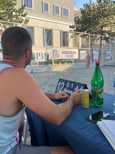 In Gošići, Montenegro, a beach volleyball match is in progress. The image focuses on a table set up near the court, with a scoreboard displaying "12" and "11". On the table, there's a green bottle of "Knjaz Miloš" mineral water, a yellow energy drink can, and a smartphone. In the background, a woman in athletic wear is playing volleyball. Several banners are visible, including one that reads "BEACHVOLLEY BEOGRAD". The scene appears to be an outdoor sporting event, likely during daylight hours.
