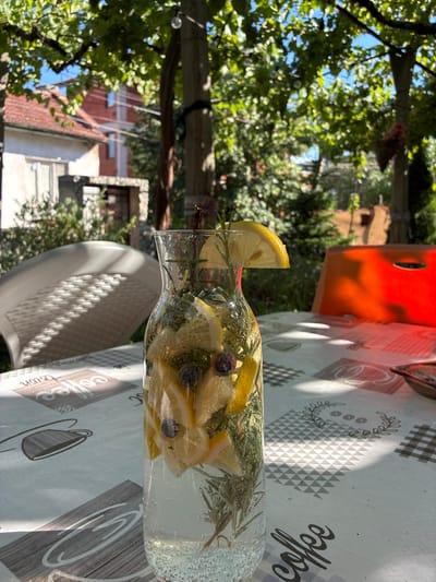 In an outdoor setting, possibly a backyard or patio, a glass carafe filled with a clear beverage and infused with lemon slices, blueberries, and rosemary sprigs sits on a patterned tablecloth. The tablecloth features coffee-themed graphics and a checkered border. Behind the carafe, a white plastic chair with a textured seat is partially visible. The background reveals a residential area with houses and lush greenery, under a bright sky. The presence of a light string with bulbs suggests an evening or garden ambiance. The scene appears to be in Simitli, Bulgaria, given the context of the request, with a focus on a refreshing drink in a natural, outdoor environment.